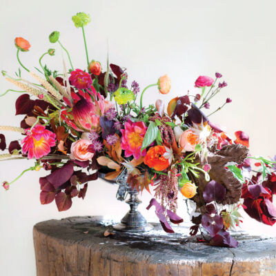 S-curve floral design in a compote vase, featuring autumnal hues along with brighter pops of pink and green