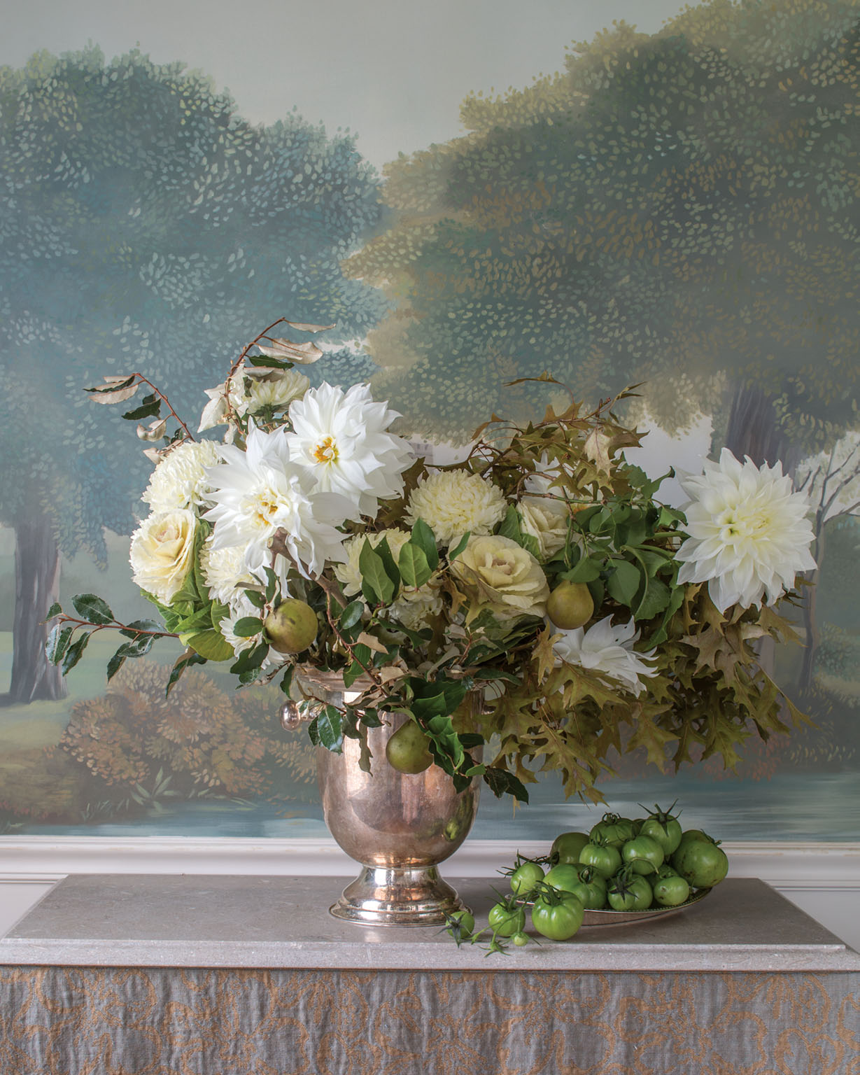 white and green floral arrangement by Sidra Forman in a silver footed vessel against the backdrop of a scenic wallpaper featuring trees