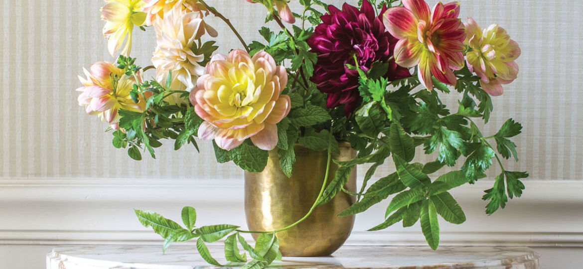 Sidra Forman's arrangement of autumn-hued dahlias with trailing herbs in a brass vessel, on a marble top occasional table with carved wood base, against a wallpaper of small taupe and white vertical stripes