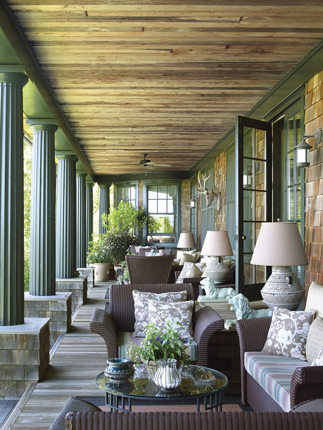 a furnished covered porch lines the side of a Hamptons home. The columns and trim are muted teal. The panel ceiling and floor are natural wood.