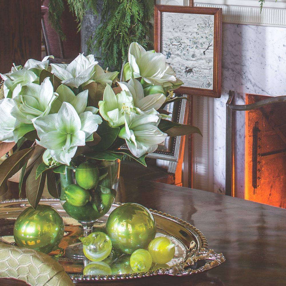 Green-centered, white amaryllis flowers arranged with magnolia leaves and limes in clear goblet.