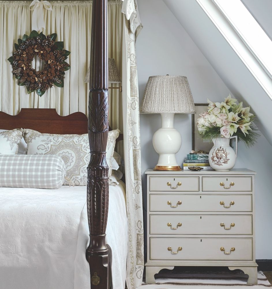 In the primary bedroom of the Williamsburg designer-in-residence house, a large pitcher of pink roses and amaryllis with pine boughs sit on the bedside table. A wreath hangs over a simple white bed.