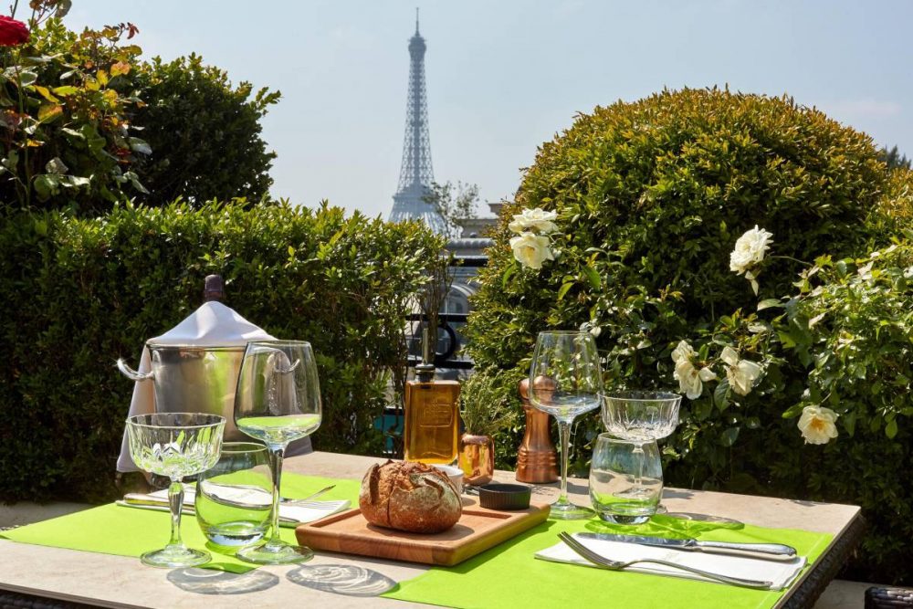 A rooftop cafe table surrounded flowering and green shrubs, with a view of the Eiffel Tower in the distance