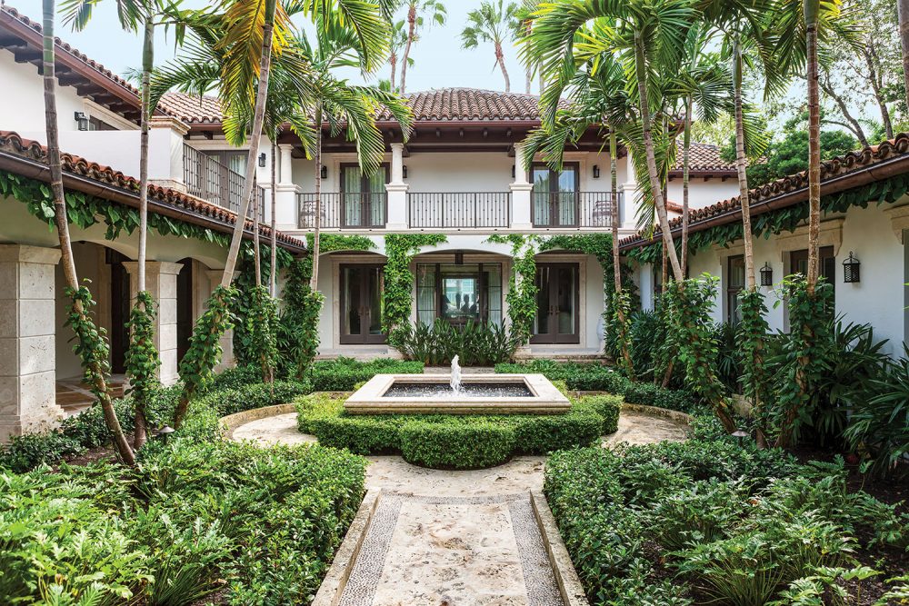Photo of a courtyard garden designed by Fernando Wong. It is surrounded on three sides by a two-story white stucco structure with a tile roof. A covered patio with columns wraps around the left and back side. The garden features ferns, palm trees and climbing greener. The limestone path leads to a circular patio with a square fountain at the center, which is surrounded by a low cotoneaster hedge.