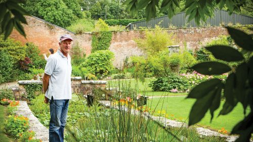 Wearing a baseball cap, polo shirt and jeans, Arthur Shackleton stands on the stone edge of a plant filled garden pond at at Dromoland Castle, holding his hands behind his back.