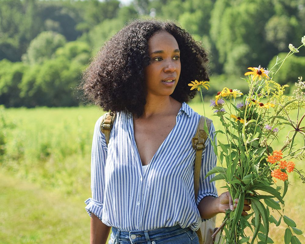 The artist stands in a green field dressed in jeans and a blue-and-white stripe blouse and a backpack.