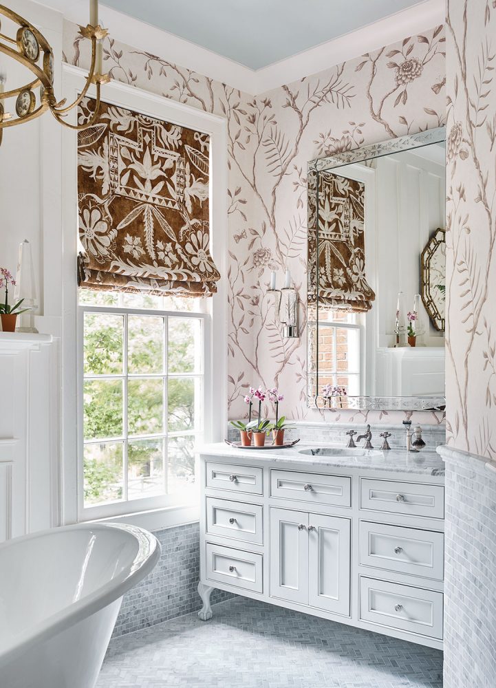 Another view of a master bath designed by James Farmer, featuring a furniture-like vanity painted white and a brown and white botanical-print Roman shade that complements the wallpaper.