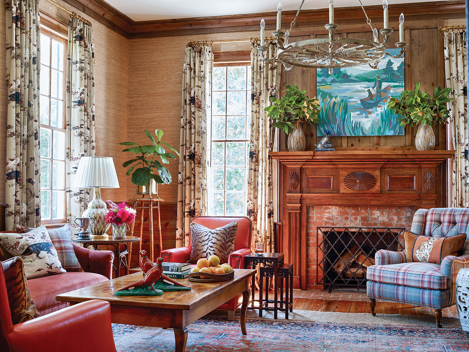 Photo of the Martin family's study designed by James Farmer. Red leather chairs, a plaid armchair and a painting featuring blues and greens bring color to the room, which features a neutral grass wallcovering, natural heart pine paneling and a brick fireplace with antique natural-wood wood mantel. A threadbare Oriental rug ties the room together.