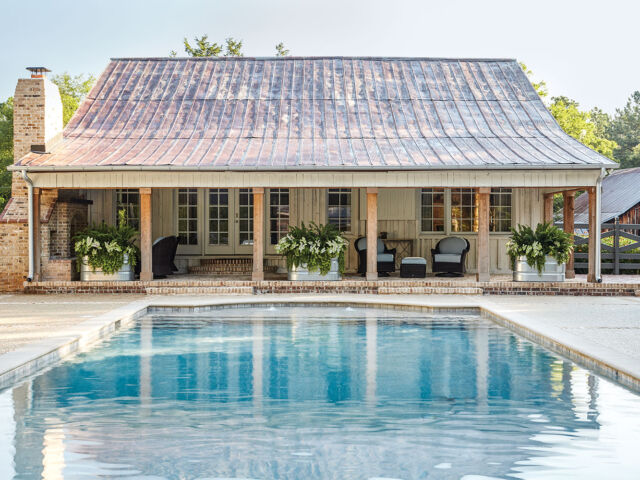 A blue pool leads to a covered brick patio of the pool house, which features wood columns across the front and an aged metal roof and brick chimney