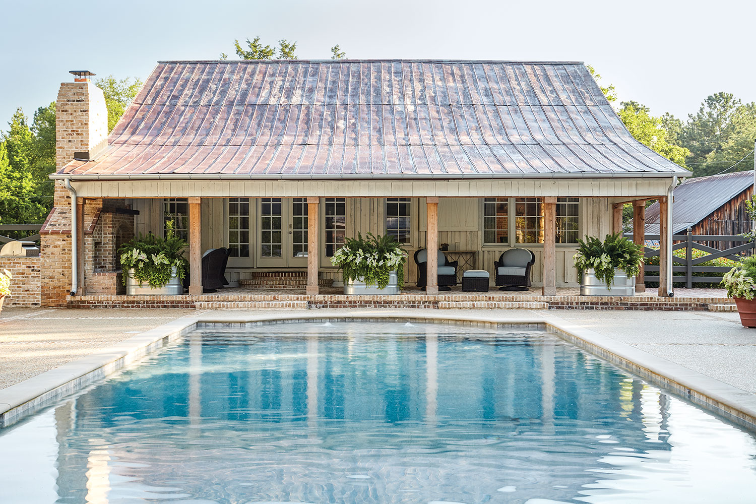 A large blue pool leads the covered brick porch the pool house, which features a weathered metal roof