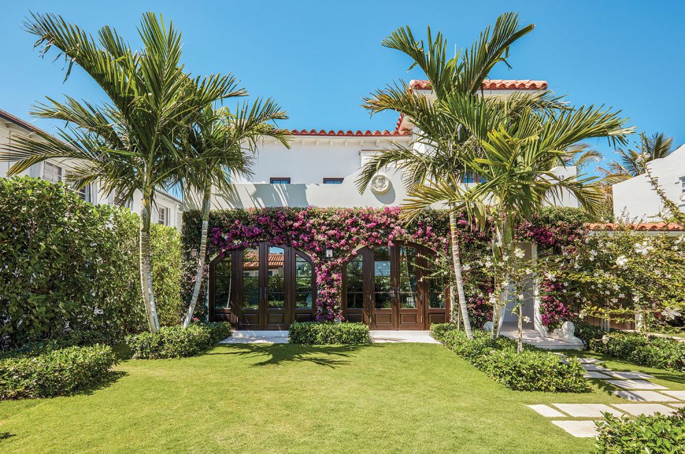 Photo of white stucco home with tile roof. The garden designed by Fernando Wong features a an expanse of open green lawn and palm trees. The first story of the side of the house facing the garden is covered in blooming bougainvillea.