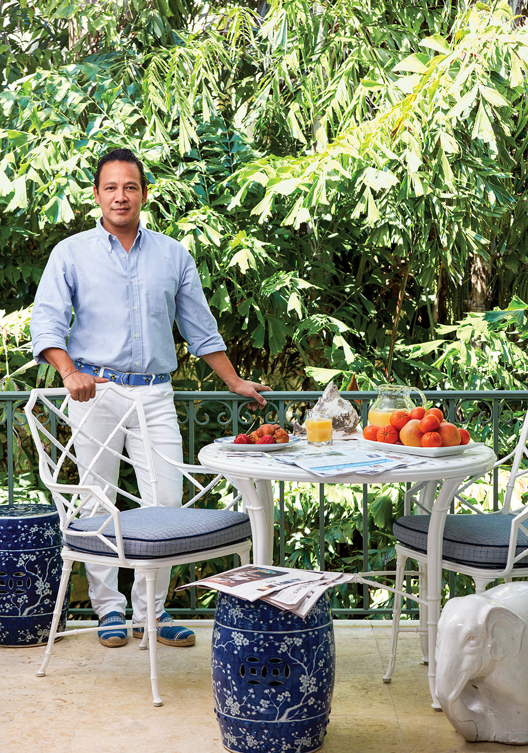 Photo of a Fernando Wong wearing white pants, a light blue shirt, and a deep blue belt and shoes. He is standing at a set of white wrought iron table and chairs.