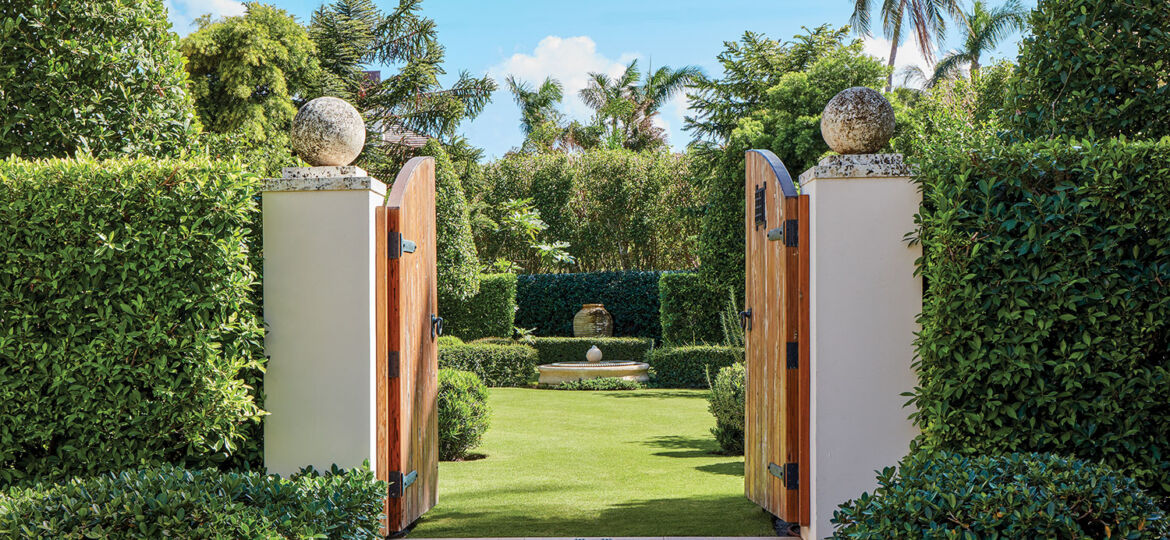 Photo of a Palm Beach Garden designed by Fernando Wong, surrounded by a high hedge. The heavy wood gate doors are open, lending a view inside of the manicured lawn, hedges and in the background a large urn.