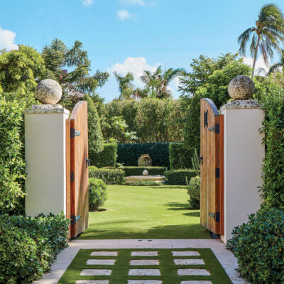 Photo of a Palm Beach Garden designed by Fernando Wong, surrounded by a high hedge. The heavy wood gate doors are open, lending a view inside of the manicured lawn, hedges and in the background a large urn.