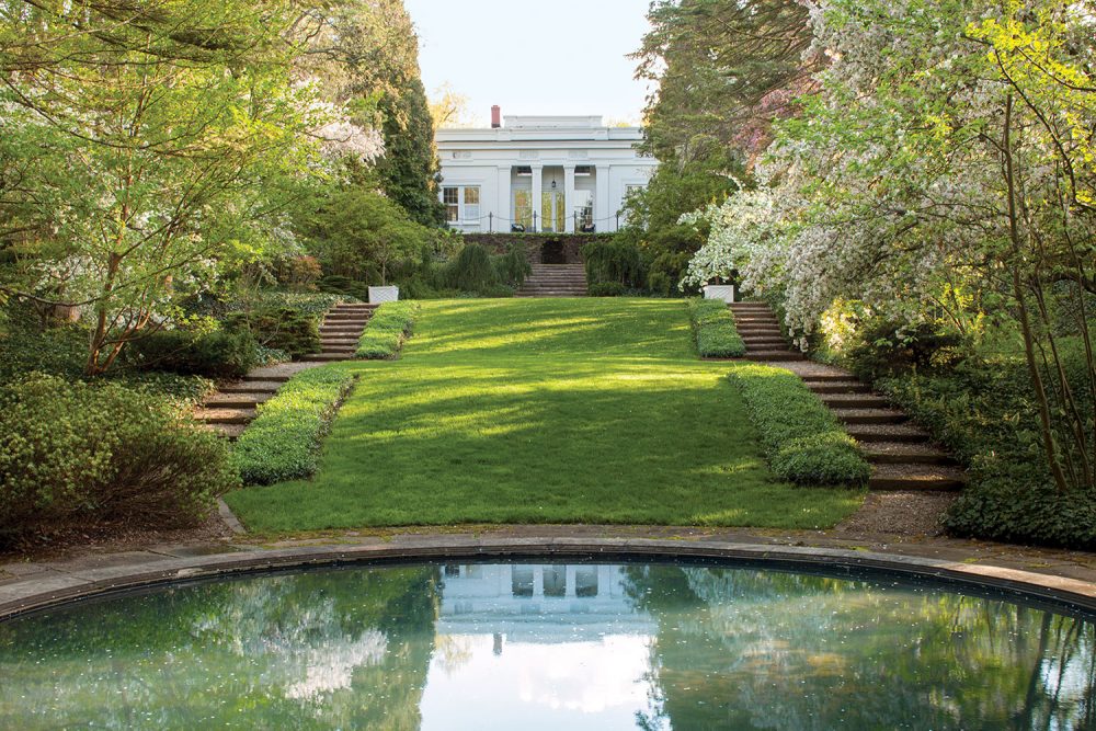 Melissa McGrain's Greek Revival home overlooks a lawn and circular pond. The historic Fletcher Steel landscape design features woodlands and a set of terraced stairs on either side of the house and lawn.
