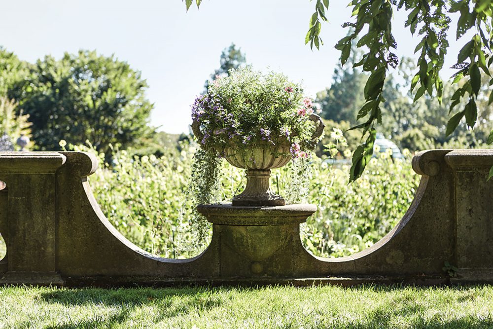 The photo shows one of the three Pennoyer Newman urns overflows with plants. The wall dips low on either side of the urn, giving a peek inside the garden.