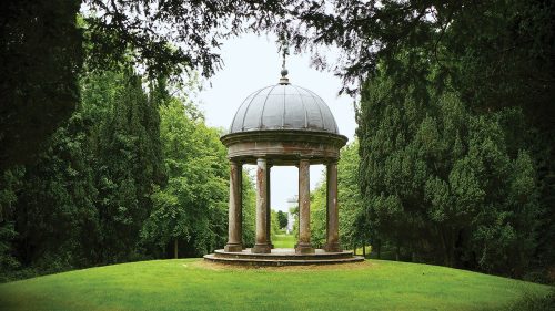 The Temple of Mercury in Ireland features a dome roof and a circular colonnade. It sits on a low mound of bright green lawn. Lush trees surround it.