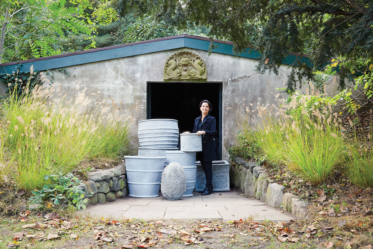 Newman stands in the doorway of a stuccoed shed with a collection of large planters. Trees and tall grasses surround the area.
