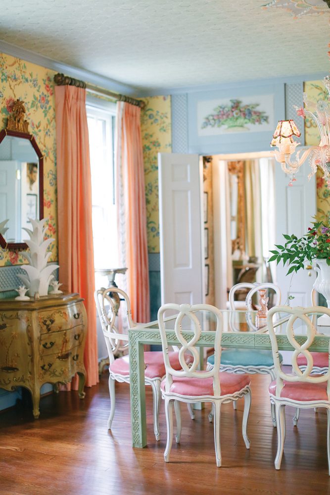 John Loecke and Jason Oliver Nixon's dining room features yellow wallpaper bearing a botanical print, a glass-top table with a carved wooden base painted light green, white painted chairs with pink upholstered seats, and peach floor-to-ceiling curtain panels