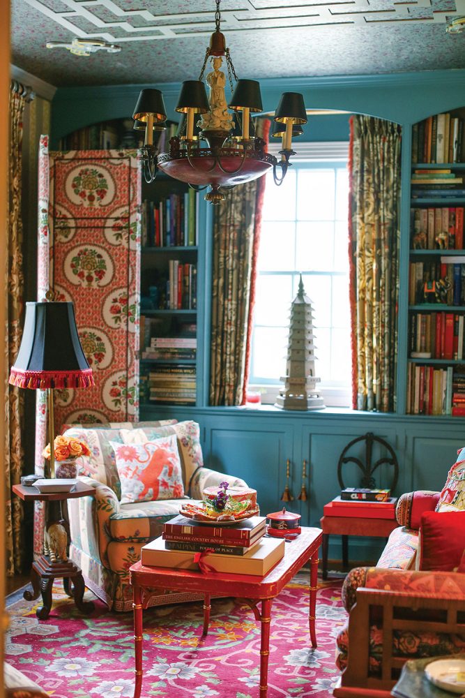The cozy Asian-inspired den features a bright oriental rug, a red laquor coffee table, upholstery boasting rich hues and patterns, and wall bookshelf/cabinetry painted a dusty blue. A small pagoda tower sits in the window.