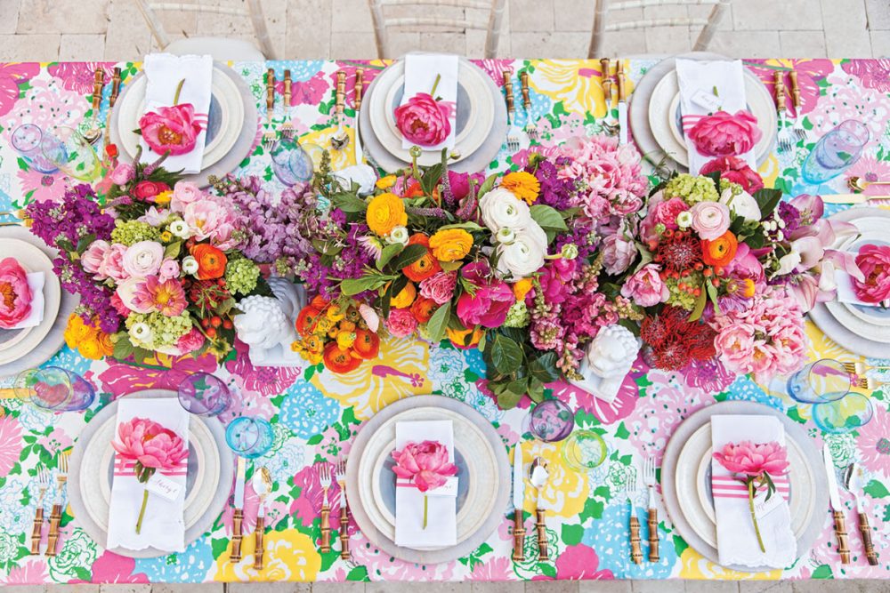 The long table is set with bamboo-handled flatware, simple white and blue plates, white scallop-edged napkins with a band of 3 pink stripes, and a pink peony atop each setting.