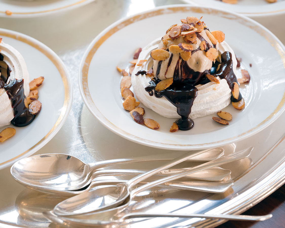 A tray of plated meringues topped with ice cream, chocolate sauce, and nuts, with silver spoons to the side, a recipe by Julia Reed