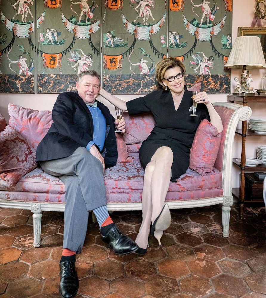 Julia Reed and Patrick Dunne sit cross-legged and sipping champagne on a pink-upholstered, wood-frame sofa in Dunne's New Orleans home