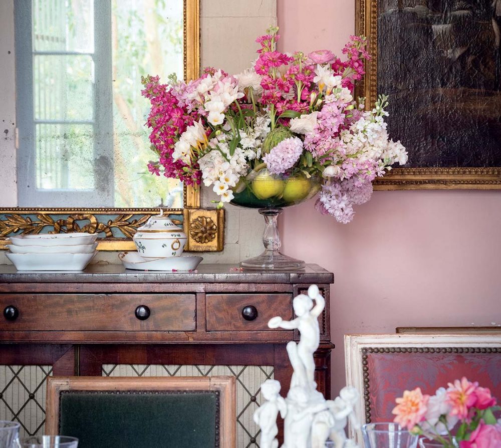A sideboard displays a large floral arrangement of pink, white and pale purple blooms in a glass pedestal bowl. Apples placed in the bowl mask the stems.