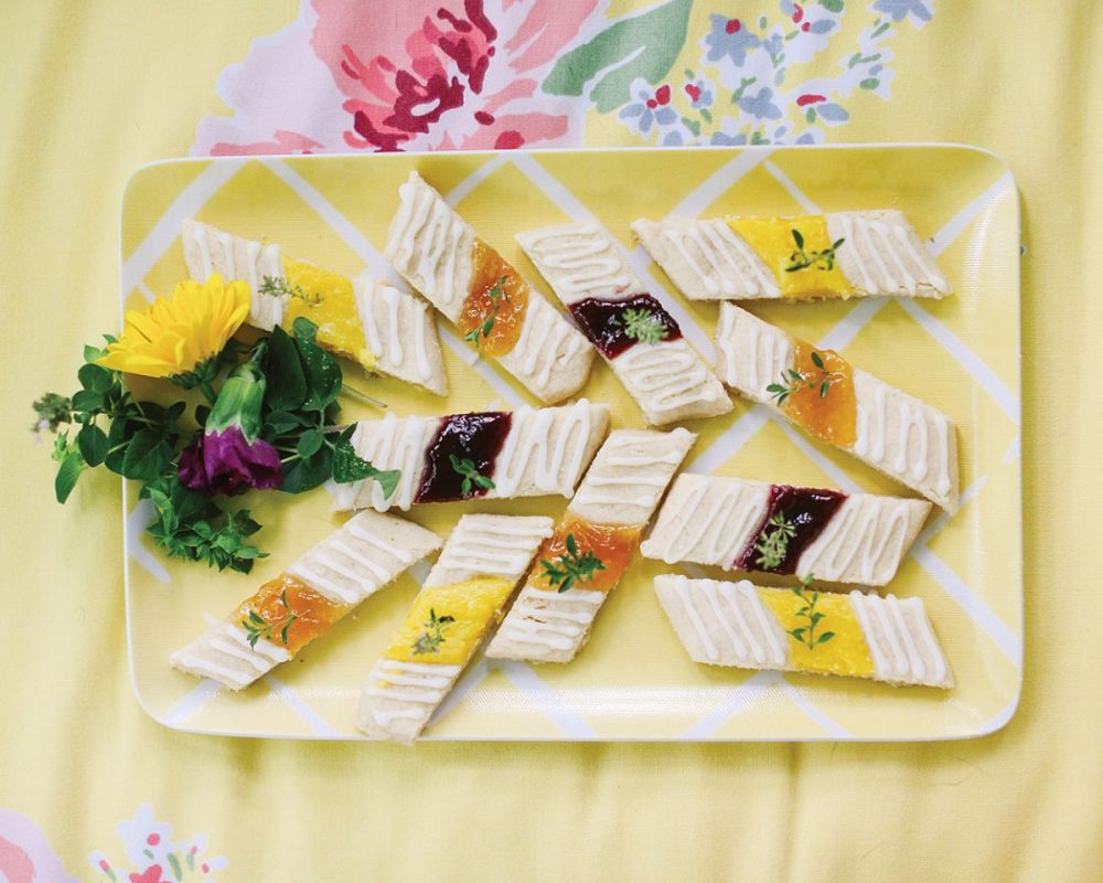 A flower-garnished platter of shortbread bars topped with various fruit fillings in the center and drizzled with a sugary glaze