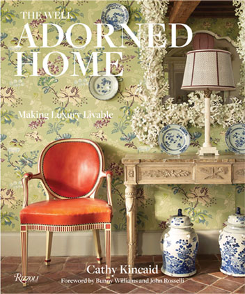 Book cover for The Well Adorned Home by Cathy Kincaid (Rizzoli New York, 2019), $50, featuring a bright orange delicate antique chair, a soothing green botanical wallpaper, entry table and mirror, and blue-and-white porcelain.