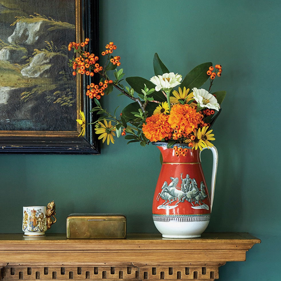 Red and white vase filled with zinnias, marigolds, black-eyed-Susans, and pyracantha on wooden table against green wall.