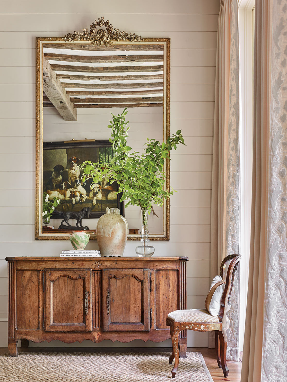 Other chic mountain house decorating ideas from this scene include a non-fussy French country-style natural wood console beneath the mirror. One top of the console are two rustic pottery jugs—one large and one small—a small stack of books, and a simple arrangement of green branches in a clear glass vase with an interesting shape. An antique French chair sits by the window, framed by soft white floor-to-ceiling curtains in curtains in a white-on-white, raised botanical pattern fabric