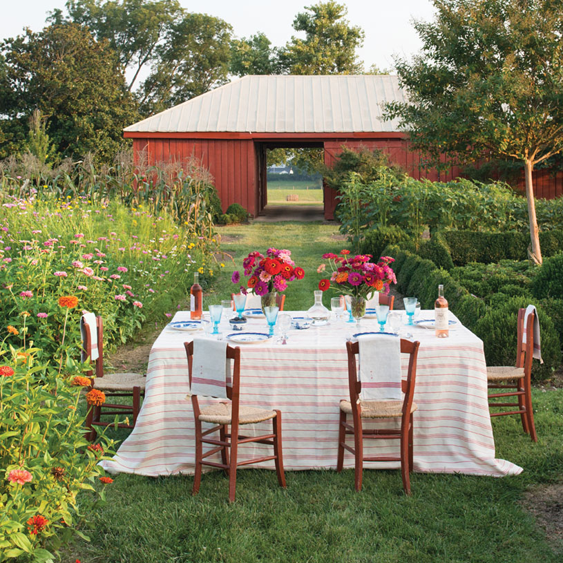 Table for six, set in a field of zinnias with a red barn in the background. The table is set with red and white linens, blue and white china, and two vases of pink and red zinnias.