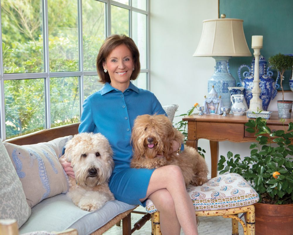In the photo, Cathy Kincaid wears a simple, collared bright blue dress. She sits on a cushioned wooden bench beside a mullioned window with her dogs on her lap. To the side, a wood side table holds a collection of blue-and-white porcelain, including the lamp.