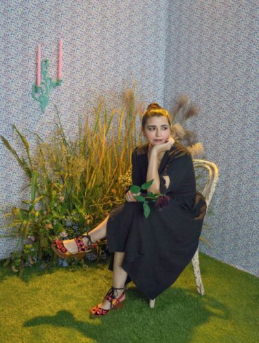 Wearing a long black dress and holding a deep burgundy rose, Marisa Bosquez-White, sits in a chair, legs crossed, resting her chin on one hand. The room features astro turf as carpet, a multicolor abstract wallpaper, and a grassy floral display. Photo by Amanda Rowan