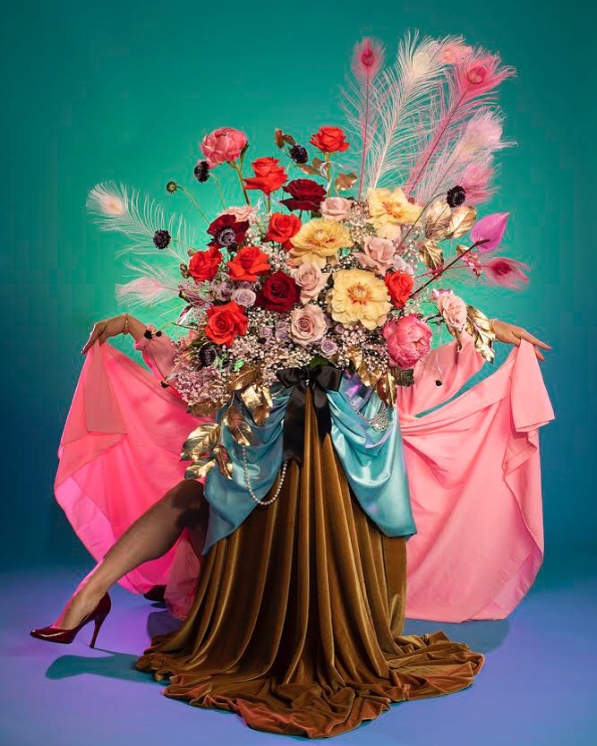 A woman's leg and arms peek out behind layers of skirts and a vibrant flower arrangement of red, purple, and yellow blooms. The scene is a collaboration of Marisa Basquez-White's floral design and Amanda Rowan's photography