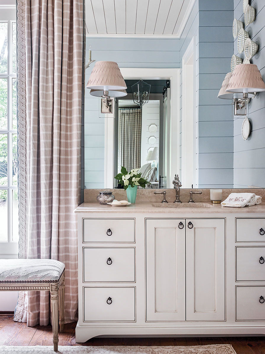 Light blue paint distinguishes the shiplap walls of the master bath from the other rooms, while white and neutral gingham curtain panels and a soft white sink vanity with a light neutral stone countertop maintain consistency with the materials Francie Hargrove used throughout her "chic mountain house" design theme.