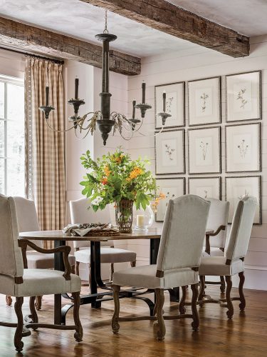 The dining room’s ivory walls and slipcovered mouton-leg chairs downplay the deep charcoal color on the chandelier and industrial steel-base table. Delicate pressed botanicals temper the bulk of the reclaimed ceiling beams. Other features include floor-to-ceiling curtain panels in a large gingham print in natural white and khaki. On the table is a loose, rustic floral arrangement of branches, including dogwood blooms, oak leaves, as well as a candle in a Simon Pearce hurricane. The framed pressed botanicals are hung in a large grid of 9 on one wall.