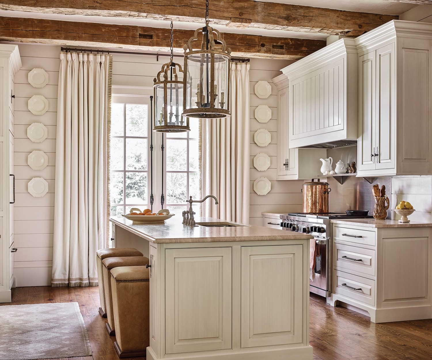 Francie Hargrove stuck with a white and neutral color palette in this mountain house kitchen, including a soft white painted finish for the cabinets and a light, neutral stone countertop. At the kitchen island, square barstools feature solid bases, simple lines, light leather upholstery, and a border of grommets just below the seat. Two large lanterns hang above the island. Five white octagonal porcelain plates hang in a vertical line on either side of soft white curtains, flanking the French doors. A large antique copper pot sits on a stainless steel oven.