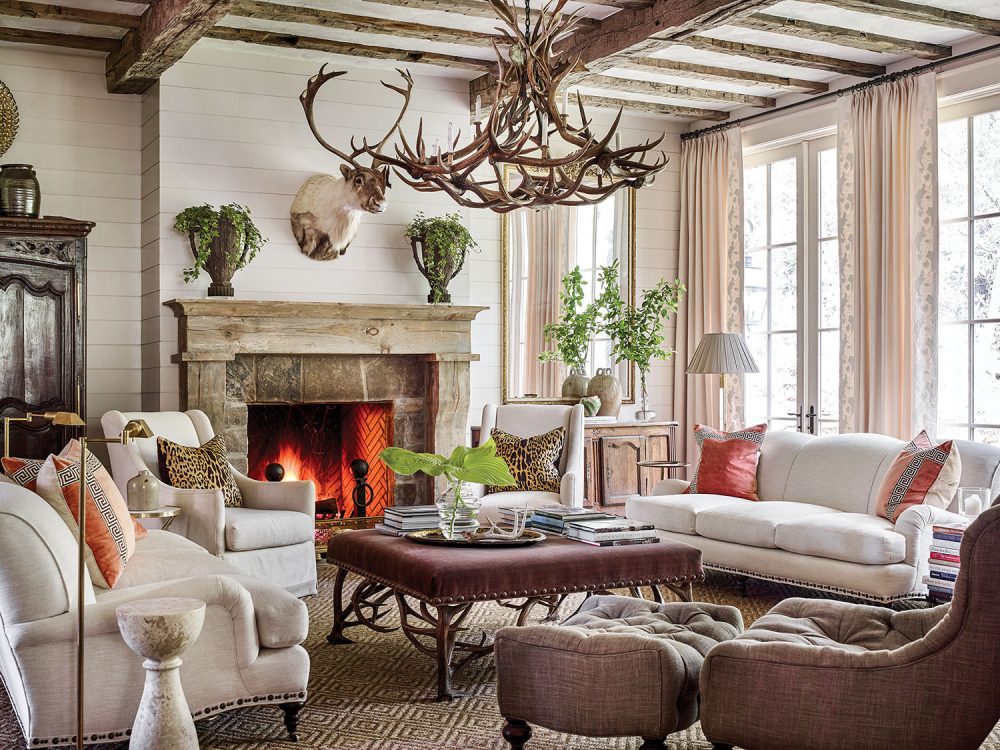 Other mountain house design ideas from this scene include a large chandelier made of deer antlers, a pair of sofas and side chairs upholstered in a soft white fabric, and throw pillows, including (on each sofa) a pair of pillows in strié coral velvet inset with a Greek key pattern, as well as (on each side chair) an leopard-print linen pillow. One tufted, curved accent chair with a matching ottoman is upholstered in a dark brown linen-type fabric. At the center of the seating area is a large square coffee table upholstered in a dark, rich fabric. A neutral patterned area rug grounds the seating area. Arrangements of simple cut greenery on the mantel, coffee table, and console add a punch of color. Swing-arm floor lamps and stacks of books invite one to sit and read by the fire. Soft white shiplap walls and curtains keep the room light and airy.