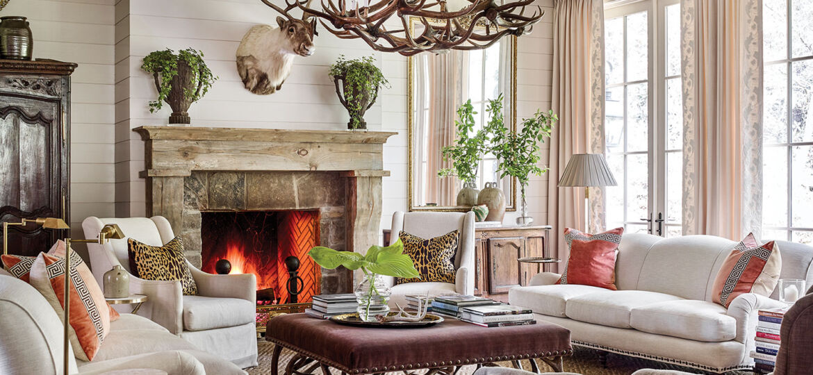 Mountain house design ideas from this scene include a large chandelier made of deer antlers, a pair of sofas and side chairs upholstered in a soft white fabric, and throw pillows, including (on each sofa) a pair of pillows in strié coral velvet inset with a Greek key pattern, as well as (on each side chair) an leopard-print linen pillow. One tufted, curved accent chair with a matching ottoman is upholstered in a dark brown linen-type fabric. At the center of the seating area is a large square coffee table upholstered in a dark, rich fabric. A neutral patterned area rug grounds the seating area. Arrangements of simple cut greenery on the mantel, coffee table, and console add a punch of color. Swing-arm floor lamps and stacks of books invite one to sit and read by the fire. Soft white shiplap walls and curtains keep the room light and airy.