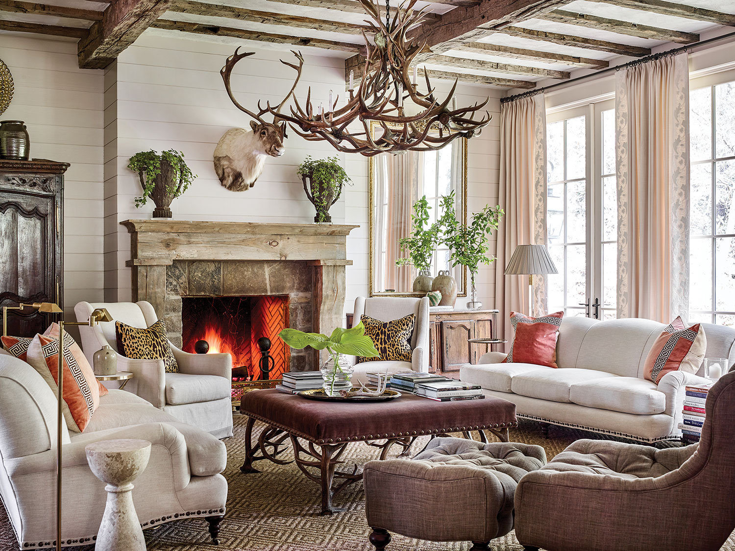 Mountain house design ideas from this scene include a large chandelier made of deer antlers, a pair of sofas and side chairs upholstered in a soft white fabric, and throw pillows, including (on each sofa) a pair of pillows in strié coral velvet inset with a Greek key pattern, as well as (on each side chair) an leopard-print linen pillow. One tufted, curved accent chair with a matching ottoman is upholstered in a dark brown linen-type fabric. At the center of the seating area is a large square coffee table upholstered in a dark, rich fabric. A neutral patterned area rug grounds the seating area. Arrangements of simple cut greenery on the mantel, coffee table, and console add a punch of color. Swing-arm floor lamps and stacks of books invite one to sit and read by the fire. Soft white shiplap walls and curtains keep the room light and airy.