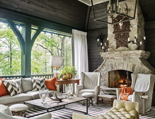 Considered to be the best seat in the house, the raised hearth of the Tennessee fieldstone fireplace makes the perfect perch for toasting marshmallows on cool evenings. A pair of candelabras carved from tree roots and an antique Belgian Black Forest plaque add to this mountain house porch’s enchanted forest vibe. Other features on this screen porch include a sofa and chairs in a light upholstery. Throw pillows and an afghan bring in a punch of bright orange that complements the muted teal millwork. A large white-and-neutral striped area rug and a pair of chevron-print throw pillows add visual interest through pattern. And though upholstered in a light fabric, a curved upholstered bench with deep, plush tufting makes a bold statement. An antique, simple wood desk holds a chunky glass lamp and a large copper pot holding a green plant. A large modern coffee table with a wood top and streamlined metal base, along with wood variety of stools and small side tables, complete the space.