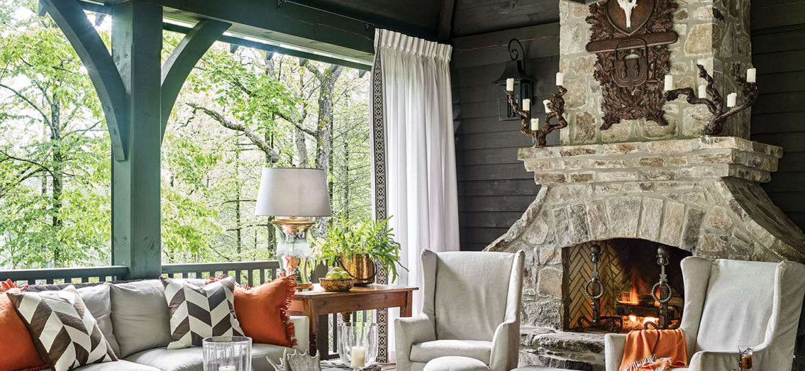 mountain house decor ideas from this screen porch include a sofa and chairs in a light upholstery. Throw pillows and an afghan bring in a punch of bright orange that complements the muted teal millwork. A large white-and-neutral striped area rug and a pair of chevron-print throw pillows add visual interest through pattern. And though upholstered in a light fabric, a curved upholstered bench with deep, plush tufting makes a bold statement. An antique, simple wood desk holds a chunky glass lamp and a large copper pot holding a green plant. A large modern coffee table with a wood top and streamlined metal base, along with wood variety of stools and small side tables, complete the space.