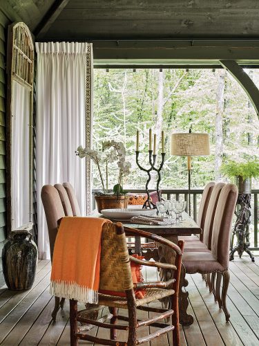 The screen porch features wood tones and other earthy hues, with a rough-hewn wooden table for 8. Francie Hargrove’s choices for mountain house decor on the porch include moth orchids in a copper pot, faux bois candlesticks and plant stand with fern, a large pottery jug with a dark glaze, an orange afghan with white fringe, a white curtain panel, and 6 leather upholstered dining chairs with grommets that compliment that pair of Appalachian hoop chairs at earth end of the table.