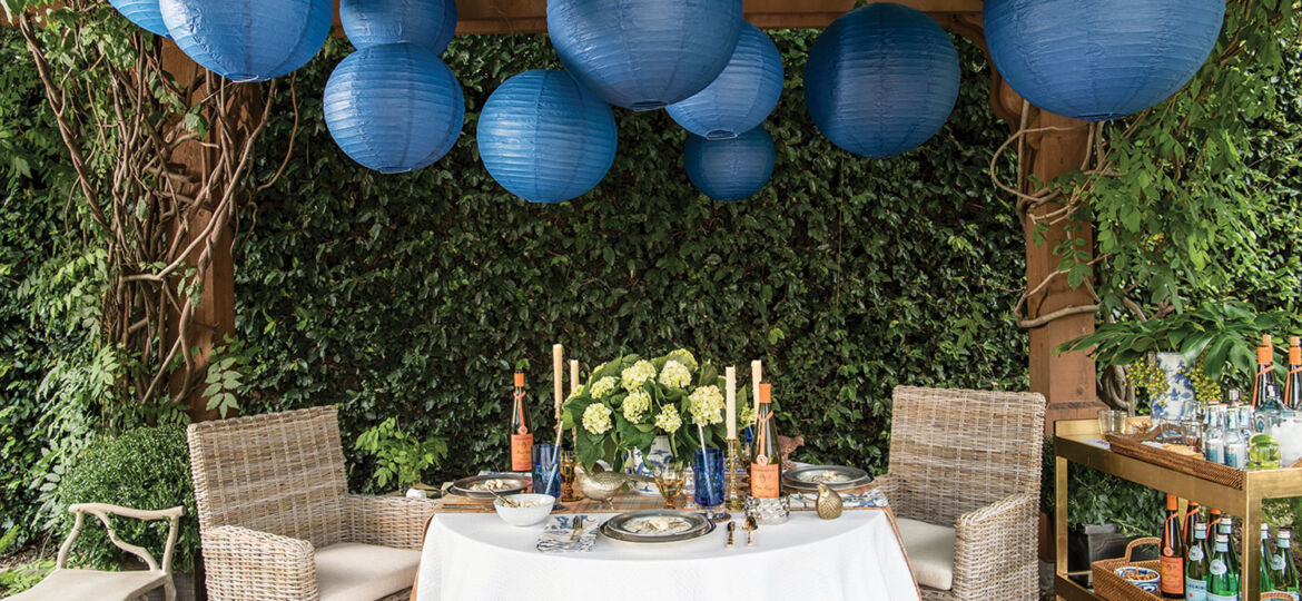 More than a dozen large, blue, spherical Chinese lanterns hang from a pergola above an outdoor party scene from the pages of Entertaining at Home by Ronda Carman. The scene also features a table with a white tablecloth and an arrangement of white hydrangeas. There are two rattan armchairs at the table. A bench sits to one side, and a stocked bar cart to the other. The background is lush with a thick wall of greenery.
