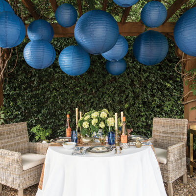 More than a dozen large, blue, spherical Chinese lanterns hang from a pergola above an outdoor party scene from the pages of Entertaining at Home by Ronda Carman. The scene also features a table with a white tablecloth and an arrangement of white hydrangeas. There are two rattan armchairs at the table. A bench sits to one side, and a stocked bar cart to the other. The background is lush with a thick wall of greenery.