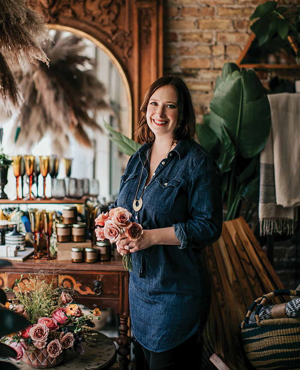 Portrait of floral designer Kelly Marie Thompson in her shop, Fleur, in Chicago. She wears a denim tunic and dark pants, and holds a small bouquet of roses. She's surrounded by flowers, grasses, large tropical leaves, and the antiques that fill the shop’s exposed brick frame.