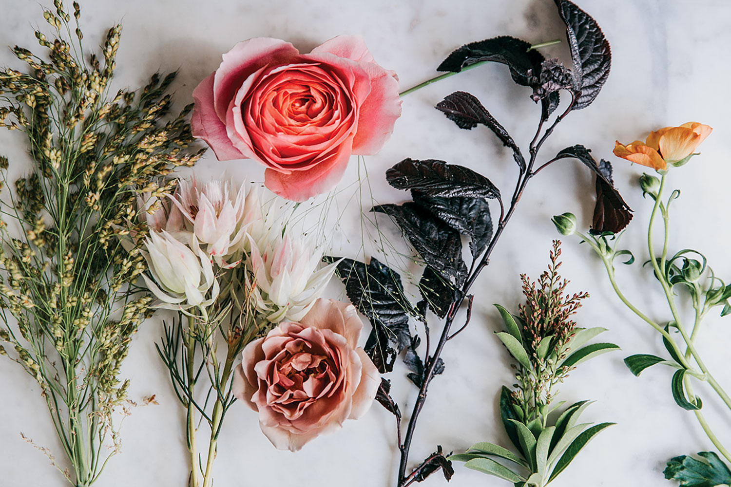 The materials are laid out on a flat white surface: Pieris japonica, Plum tree foliage, Bridal veil grass , Millet grass, Explosion grass, ‘Blushing Bride’ protea‘, Prairie Sunset’ roses, ‘Romantic Antike’ roses, ‘Koko Loko’ roses, ‘Butterfly’ ranunculus