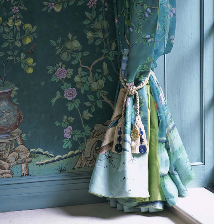 Close up of a blue-green floral curtain tied back with a tassel, which is complemented by the similarly hued wallpaper